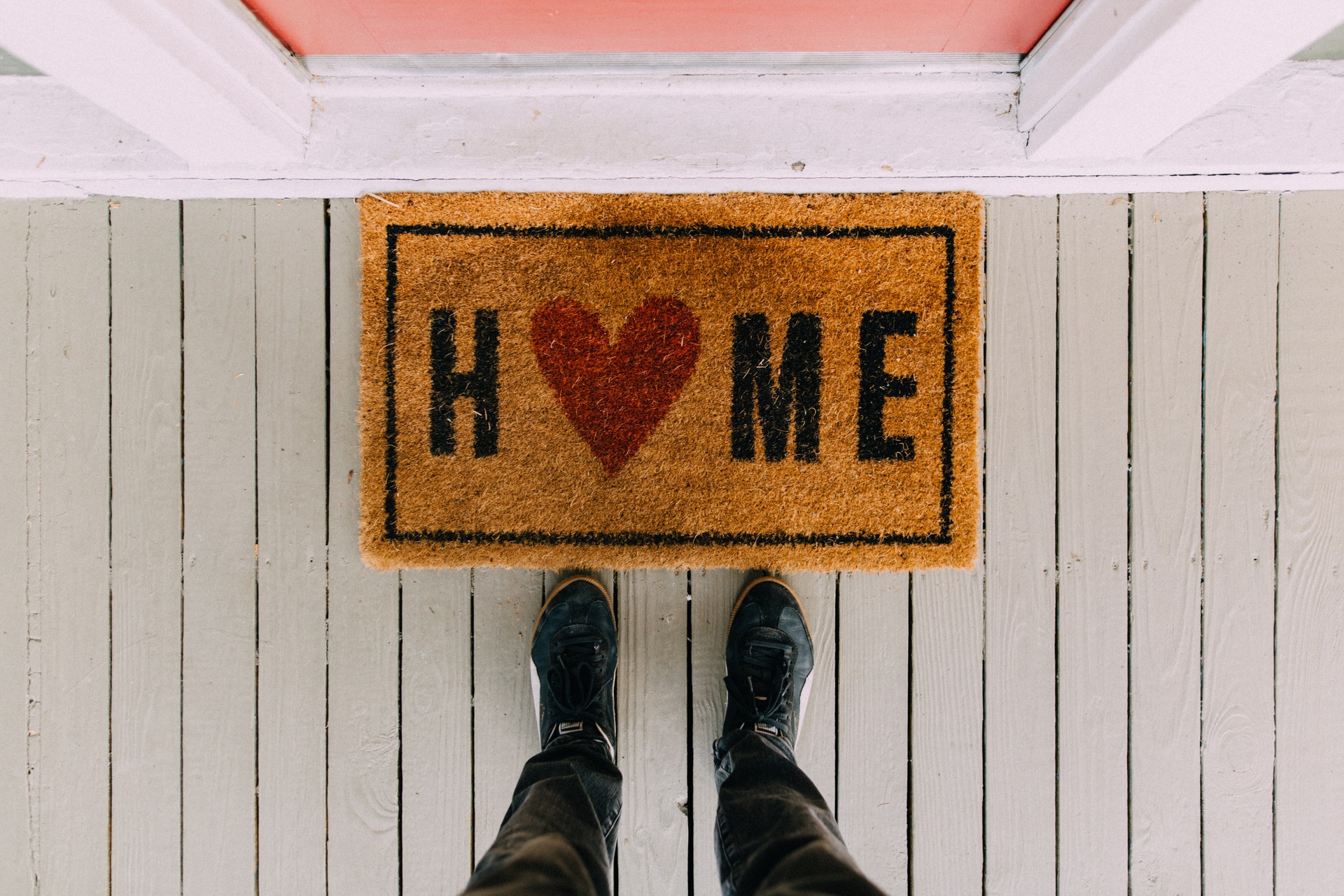 A view of a welcome mat saying HOME on it, with a man's shoes visible in front of it, as if he is taking the photo looking down
